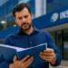 Homem de camisa azul marinho, barba grisalha, expressão preocupada, segura pasta azul com papéis diante do prédio do INSS.