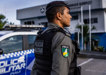 Policial da PM-PI com colete tático preto e boina observa área externa em frente a viatura azul e branca.