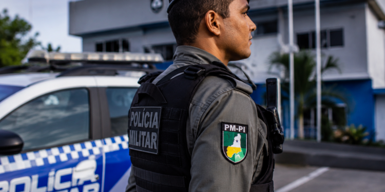 Policial da PM-PI com colete tático preto e boina observa área externa em frente a viatura azul e branca.
