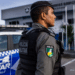Policial da PM-PI com colete tático preto e boina observa área externa em frente a viatura azul e branca.