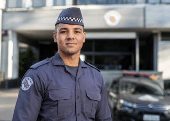 Policial militar com uniforme cinza da PMESP, quepe xadrez e brasão bordado, em frente a viatura e prédio oficial.