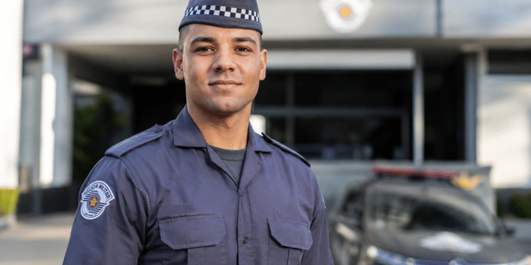 Policial militar com uniforme cinza da PMESP, quepe xadrez e brasão bordado, em frente a viatura e prédio oficial.
