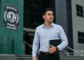 Homem de camisa azul clara segurando pastas e caderno em frente ao brasão da Polícia Penal do Maranhão.
