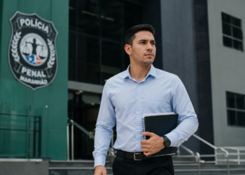 Homem de camisa azul clara segurando pastas e caderno em frente ao brasão da Polícia Penal do Maranhão.