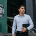 Homem de camisa azul clara segurando pastas e caderno em frente ao brasão da Polícia Penal do Maranhão.