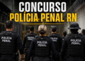 Agentes da Polícia Penal de costas com coletes táticos pretos diante de portão amarelo de unidade prisional.