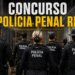 Agentes da Polícia Penal de costas com coletes táticos pretos diante de portão amarelo de unidade prisional.