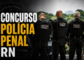 Três policiais penais de costas com uniformes pretos e coletes táticos com inscrição "Polícia Penal"