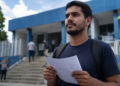 Jovem de camiseta azul-marinho segura formulário e caneta frente a escadaria de prédio público azul.