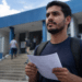Jovem de camiseta azul-marinho segura formulário e caneta frente a escadaria de prédio público azul.