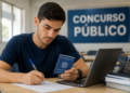 Jovem de camiseta azul preenche formulário segurando Carteira de Trabalho, com laptop e placa Concurso Público ao fundo.