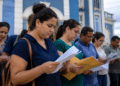 Grupo de pessoas lendo folhas e envelopes amarelos em fila externa junto a prédio colonial azul e branco.