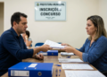 Homem de camisa azul entrega papéis a funcionária em setor de inscrições de concurso de prefeitura