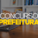 Notebook aberto sobre mesa de madeira com estante de livros ao fundo e texto "Concurso Prefeitura" em amarelo e branco.