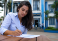 Mulher de camisa azul clara escreve em documento sobre mesa de madeira, com prédio azul institucional ao fundo.