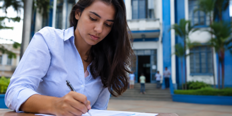 Mulher de camisa azul clara escreve em documento sobre mesa de madeira, com prédio azul institucional ao fundo.