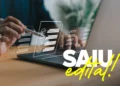 Mão segura caneta stylus sobre laptop com ícones de checklist digital e texto "SAIU edital!" em destaque.