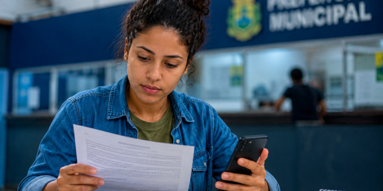 Mulher de cabelos cacheados com jaqueta jeans analisa documento segurando celular em balcão de Prefeitura Municipal.