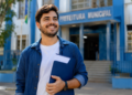 Jovem brasileiro de camisa jeans azul segura pasta azul marinho sorrindo em frente ao prédio da Prefeitura Municipal.