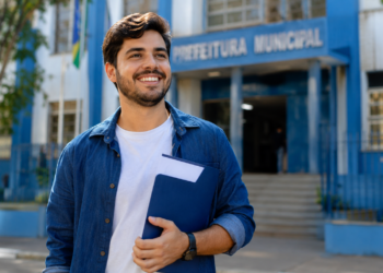 Jovem brasileiro de camisa jeans azul segura pasta azul marinho sorrindo em frente ao prédio da Prefeitura Municipal.