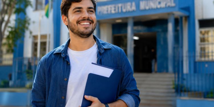 Jovem brasileiro de camisa jeans azul segura pasta azul marinho sorrindo em frente ao prédio da Prefeitura Municipal.
