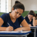 Mulher de coque alto e camiseta azul-marinho escreve em folha de papel durante prova em sala com parede verde.
