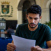 Homem de cabelos cacheados e camiseta verde escura sentado em cadeira azul lendo papel em recepção com brasão ao fundo.