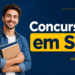 Jovem de barba curta segura cadernos, veste camisa jeans sobre camiseta branca com mochila preta nas costas