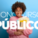 Jovem negra sorridente com cabelo afro, óculos e blusa rosa segura livros e documentos sobre fundo azul.