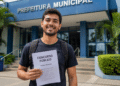 Jovem brasileiro com mochila segura caderno de prova objetiva de concurso público diante de prédio da Prefeitura Municipal.