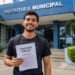 Jovem brasileiro com mochila segura caderno de prova objetiva de concurso público diante de prédio da Prefeitura Municipal.