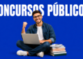 Jovem de óculos sentado com laptop no colo ergue o punho em comemoração, ao lado de livros empilhados