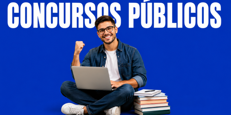 Jovem de óculos sentado com laptop no colo ergue o punho em comemoração, ao lado de livros empilhados