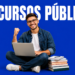Jovem de óculos sentado com laptop no colo ergue o punho em comemoração, ao lado de livros empilhados