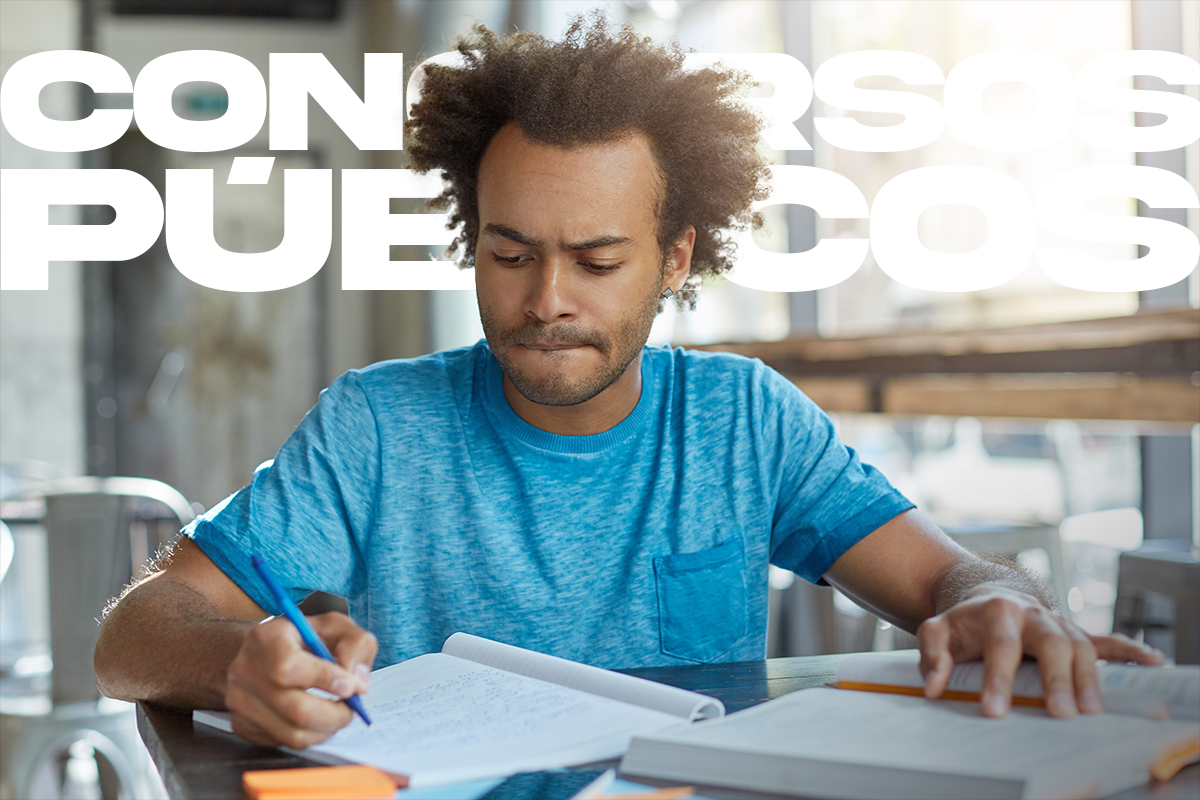 Jovem de cabelos crespos e camiseta azul escrevendo em caderno durante sessão de estudos para concurso público.