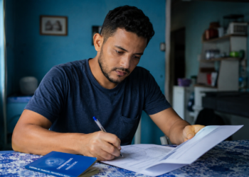 Homem de camiseta azul escreve em documentos com caneta, carteira de trabalho azul da Previdência Social visível.