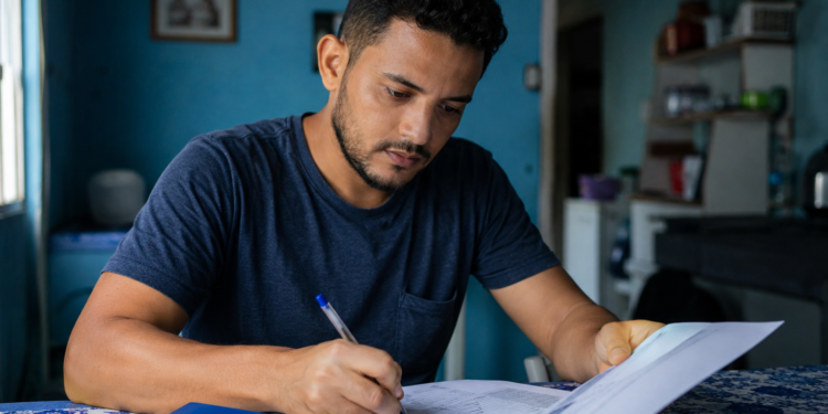 Homem de camiseta azul escreve em documentos com caneta, carteira de trabalho azul da Previdência Social visível.