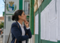 Mulher de blazer azul e camisa branca analisa lista de aprovados fixada em mural verde de prédio público.