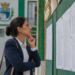 Mulher de blazer azul e camisa branca analisa lista de aprovados fixada em mural verde de prédio público.