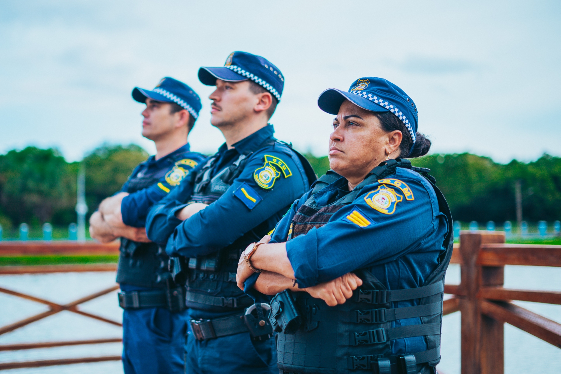 Três guardas civis municipais de braços cruzados em fila, com uniformes azuis e coletes táticos, em área de lago