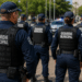 Guardas municipais de costas com coletes pretos e uniformes azul-marinho em formação, viatura policial ao fundo.