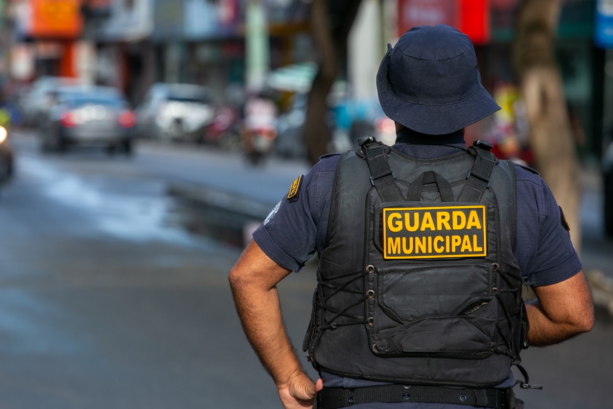 Guarda Municipal de costas com colete tático preto e boné azul observa rua comercial movimentada