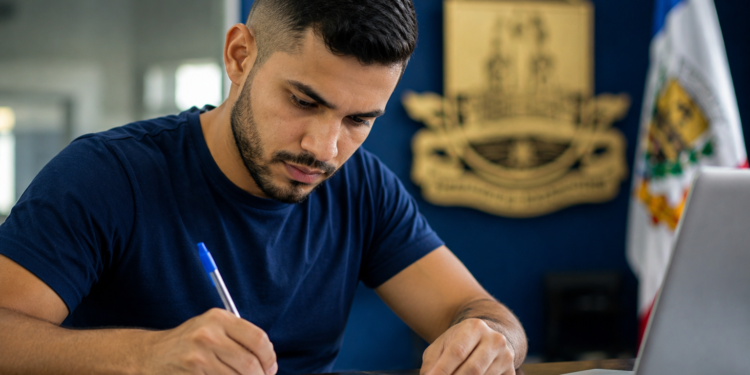 Jovem de camiseta azul-marinho escreve em documento com caneta azul, diante de notebook, em sala com brasão e bandeira de MG