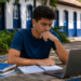 Jovem de camiseta azul estuda com laptop e cadernos em mesa externa, frente a casarão colonial azul e branco.