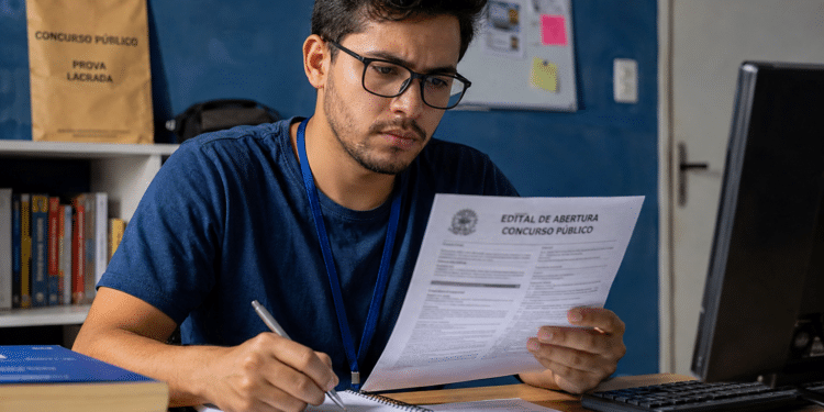 Jovem de óculos e camiseta azul analisa edital de concurso público enquanto faz anotações em caderno.