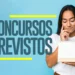 Mulher jovem de cabelo escuro com camiseta branca segura calendário enquanto apoia queixo com a mão em gesto pensativo