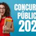 Jovem de cabelos cacheados, óculos e blusa vermelha segura livros com expressão surpresa diante de texto sobre concursos 2026