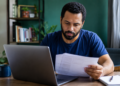 Homem de barba com camiseta azul marinho analisa papéis em frente a notebook cinza em escritório doméstico.