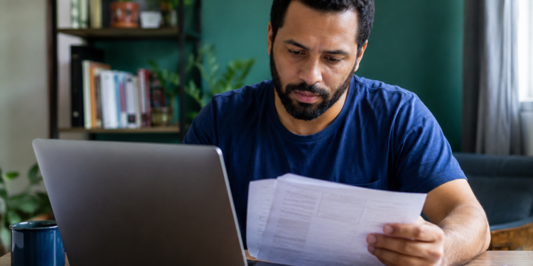 Homem de barba com camiseta azul marinho analisa papéis em frente a notebook cinza em escritório doméstico.