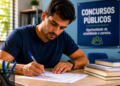 Homem jovem de camiseta azul marinho escreve com caneta em documentos ao lado de livros empilhados em ambiente de estudos.